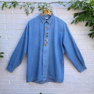 May include: A light blue long-sleeve denim shirt featuring a vertical pleated panel down the center front. The panel is decorated with a series of small, colorful embroidered motifs depicting various outdoor and golf-related scenes and figures.