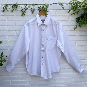 May include: A crisp white long-sleeve button-down shirt featuring a classic collar, a single chest pocket with a flap, and a central button placket. The garment includes a small white tag attached to the front buttons and long sleeves with cuffs.