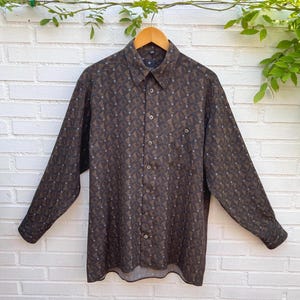 May include: A long-sleeve button-down shirt in a dark brown hue featuring a repeating geometric pattern of small, light-colored squares. The shirt includes a standard collar, a front button placket, and a single chest pocket on the left side.
