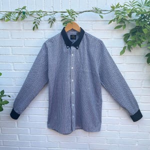 May include: A long-sleeve button-down shirt featuring a fine black and white vertical pinstripe pattern. The garment includes a solid black pointed collar, black cuffs, a front button placket with white buttons, and a single chest pocket on the left.