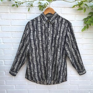 May include: A long-sleeve button-down shirt featuring a black and white vertical striped pattern with intricate geometric and floral motifs. The shirt has a standard collar, a front button placket, and buttoned cuffs, presented on a wooden hanger.