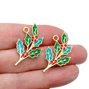 May include: Two gold-coloured holly leaf charms with green and teal enamel leaves and red berry accents. Each charm has a small loop at the top for attaching to jewellery or other crafts. The charms are held in a hand.
