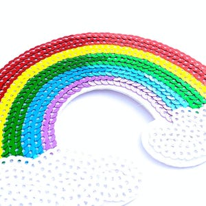 May include: A rainbow-shaped patch covered in vibrant, multi-coloured sequins arranged in arched rows of red, orange, yellow, green, blue, and purple, with white sequin clouds at both ends of the arc against a plain white background.