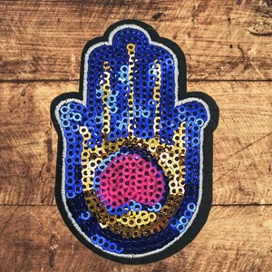 Könnte beinhalten: Ein paillettenbesetzter Hamsa-Hand-Aufnäher mit schwarzem Rand. Die Hand ist hauptsächlich blau mit goldenen und rosa Pailletten. Der Aufnäher befindet sich auf einer Holzoberfläche.