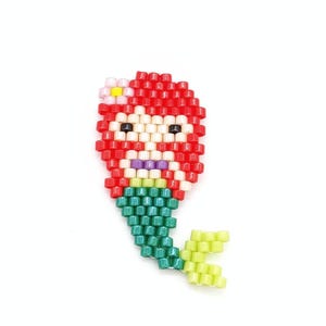 May include: A beaded pin shaped like a mermaid with bright red hair, a small white flower with a yellow centre, a pale skin tone, purple lips, and a green tail with a light yellow fin, all constructed from small, uniform, glossy plastic beads.