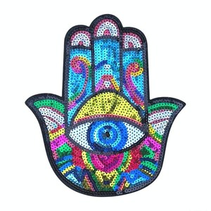 May include: A vibrant sequined Hamsa hand patch with an eye motif. The patch has a black outline and is embellished with blue, yellow, pink, and green sequins. The eye, with a blue iris and yellow details, is centrally positioned.