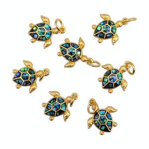 May include: Gold-coloured turtle charms with iridescent blue, green, and black shell inlays. The charms have a textured finish and small loops for attaching to jewellery. Several charms are displayed against a white background.
