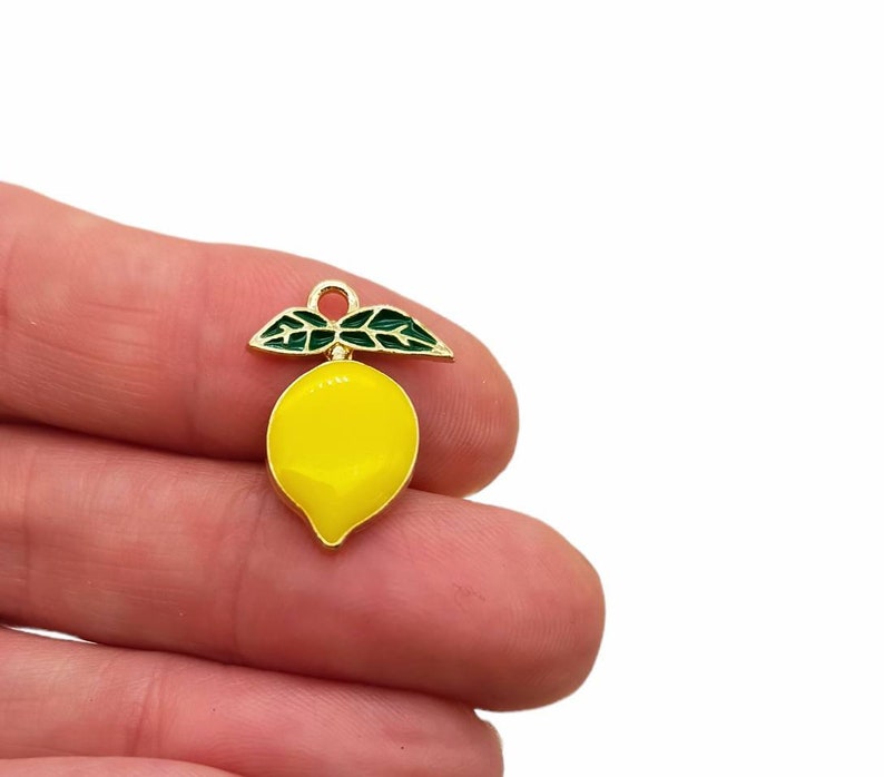 Puede incluir: Un peque&ntilde;o dije con forma de lim&oacute;n que presenta un cuerpo de esmalte amarillo brillante con un borde de metal dorado. En la parte superior, tiene dos hojas de esmalte verde oscuro y un peque&ntilde;o aro de metal dorado para colgar.
