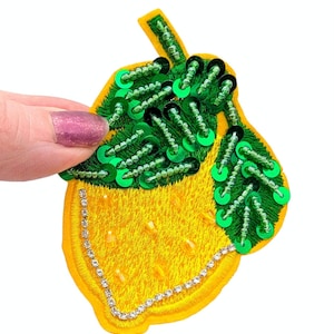 May include: Embroidered lemon patch with yellow fabric, green sequined leaves, and a rhinestone border. The patch is held by a hand with pink nail polish. The lemon design is a bright, cheerful embellishment.