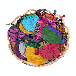 May include: A collection of small, round, drawstring pouches made from various colourful patterned fabrics, including shades of yellow, pink, green, blue, and purple, all arranged together inside a round, woven, light-coloured wicker basket.