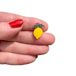 Puede incluir: Un peque&ntilde;o dije con forma de lim&oacute;n que presenta un cuerpo de esmalte amarillo brillante y dos hojas de esmalte verde oscuro en la parte superior. El dije est&aacute; delineado en metal dorado e incluye una peque&ntilde;a argolla circular en la parte superior.