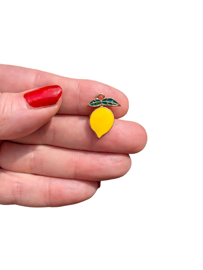 Puede incluir: Un peque&ntilde;o dije con forma de lim&oacute;n que presenta un cuerpo de esmalte amarillo brillante y dos hojas de esmalte verde oscuro en la parte superior. El dije est&aacute; delineado en metal dorado e incluye una peque&ntilde;a argolla circular en la parte superior.