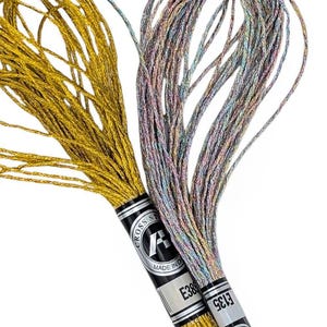 May include: Two spools of metallic embroidery thread. One is gold, the other a blend of silver, pink, and blue. The spools are labelled with the brand name and product codes. Ideal for cross stitch and embroidery projects.