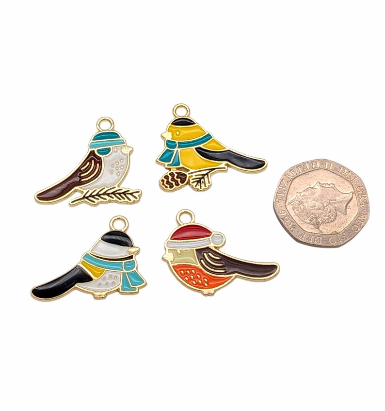 May include: Four gold-tone metal charms shaped like birds wearing winter accessories. The birds are painted in various colors, including red, yellow, blue, white, and black. The charms are approximately the size of a British one pound coin.
