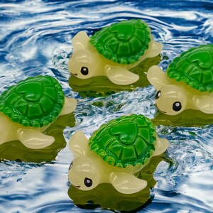 May include: Four small, yellow, plastic toy turtles with green shells are floating in blue water. The turtles have black eyes and a heart-shaped design on their shells.