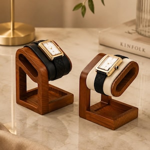 May include: Two rectangular wristwatches with gold-tone cases and white faces, each featuring a black textured leather strap. The watches are displayed on individual C-shaped wooden stands with a natural brown finish and a square base.