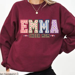 Può includere: Una felpa girocollo bordeaux con il nome EMMA in grandi lettere stampate a motivi vari. Ogni lettera presenta un design unico, tra cui quadretti vichy rossi, fiori, strisce blu e pois viola. Sotto è stampata la scritta CHEER MOM.