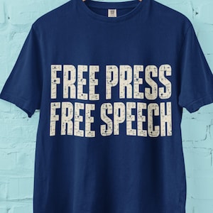 May include: A navy blue short-sleeved t-shirt featuring the text "FREE PRESS FREE SPEECH" printed in a distressed, off-white, block-style font across the chest. The garment has a crew neckline and is displayed on a wooden hanger.