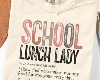 May include: An ivory t-shirt featuring the text SCHOOL LUNCH LADY in muted pink and brown tones. Below, the dictionary-style definition reads: noun /skool lən(t)sh 'lādē/ Like a chef who makes yummy food for everyone every day. See also: mealtime magicians, food friends.