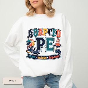 May include: A white long-sleeve sweatshirt featuring a graphic design with the text ADAPTED PE. The design includes a colorful ball, a blue sneaker, an orange traffic cone, and a blue whistle. Below, a banner displays the words Move, Include, Empower.