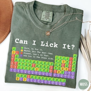Op de afbeelding: Een olijfgroen t-shirt met de witte tekst Can I Lick It? boven een kleurrijke grafiek van het periodiek systeem. De tabel bevat een legenda met vier kleurgecodeerde veiligheidsniveaus variërend van groen tot paars, gedrukt op een shirt met ronde hals.
