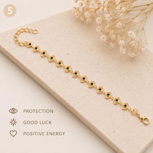 Pode incluir: Uma pulseira de corrente em tom dourado com uma série de elos repetidos em forma de olho, cada um centrado com uma pequena conta preta. A pulseira inclui um fecho lagosta e uma corrente extensora. O texto na imagem diz: PROTECTION, GOOD LUCK, POSITIVE ENERGY.