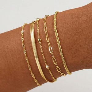 May include: A set of five gold-toned bracelets featuring varied chain styles, including a twisted rope chain, a flat herringbone chain, a delicate cable chain with small round beads, an elongated oval link chain, and a second twisted rope chain.