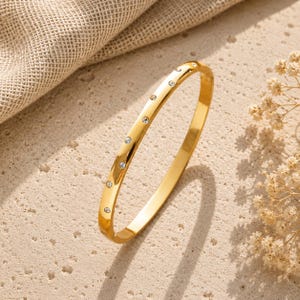May include: A polished gold-tone bangle bracelet featuring a series of small, clear, round-cut stones embedded along the outer surface. The metal has a smooth, reflective finish, and the stones are spaced evenly in a single row along the band.