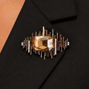 May include: A brooch featuring a large, rectangular, champagne-colored central crystal surrounded by a sunburst arrangement of thin gold-tone metal bars, accented with small white pearls, deep red stones, and tiny blue crystals.