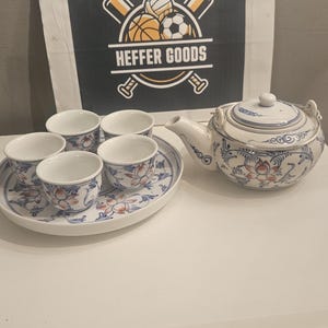 Könnte beinhalten: Ein weißes Keramik-Teeservice bestehend aus einer Teekanne, vier kleinen Tassen und einem runden Tablett, alle mit blau-orangen Blumenmustern verziert. Dahinter steht ein rechteckiges Schild mit dem Text HEFFER GOODS und Sportball-Symbolen.