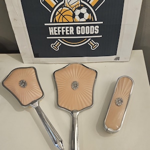 Könnte beinhalten: Ein Vintage-Toilettenset bestehend aus einem Handspiegel, einer Haarbürste und einer Kleiderbürste. Jedes Teil hat eine blasspfirsichfarbene Rückseite mit Strahlenmuster, einen silberfarbenen Metallrahmen und ein florales Emblem. Schild: HEFFER GOODS.
