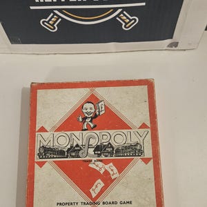 May include: A vintage Monopoly property trading board game box with a red and cream color scheme. The cover features a cartoon character holding a pound note, a train illustration, and text reading Monopoly, John Waddington Ltd, London & Leeds.