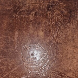 May include: A brown leather surface featuring a detailed embossed portrait of a soldier wearing a tall bearskin hat and a military uniform with braided epaulets and buttons. The leather has a textured, weathered appearance with visible stitching.