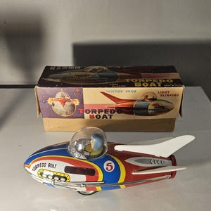 Könnte beinhalten: Ein Torpedoboot-Spielzeug aus Blech im Vintage-Stil mit einer rot-blau-weißen Farbgebung, einer transparenten Kunststoff-Cockpitkuppel, einer Pilotenfigur und der Nummer 5 an der Seite, präsentiert vor der dazugehörigen Pappschachtel.