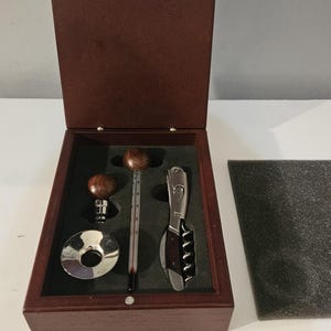 May include: A dark brown wooden box containing a wine accessory set. The set includes a silver-coloured bottle stopper with a round wooden top, a silver-coloured drip ring, a glass thermometer with a wooden top, and a folding corkscrew with wood accents.