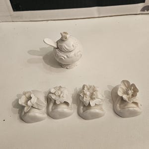 Könnte beinhalten: Ein weißes Keramik-Gewürzgefäß mit Deckel und kleinem Löffel, begleitet von vier einzelnen weißen Keramik-Blumenornamenten. Die Gegenstände sind vor einer schwarz-weißen Schachtel mit der Aufschrift HEFFER GOODS platziert.