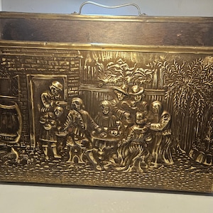 May include: A rectangular brass magazine rack featuring a detailed embossed relief scene of a group of people gathered around a table outdoors. The container has a metal handle at the top and a dark interior, with a textured brick wall and tree.