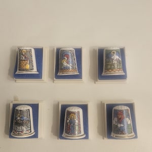 May include: A collection of six ceramic thimbles, each featuring a colourful stained-glass style religious illustration. The thimbles are individually displayed in small, square white plastic cases with blue velvet-like inserts for secure storage.