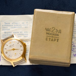May include: A gold-coloured vintage wristwatch with a white face, black numerals, and a subdial. Beside it is a small, rectangular, tan box with Cyrillic text and a paper with Cyrillic text and stamps.