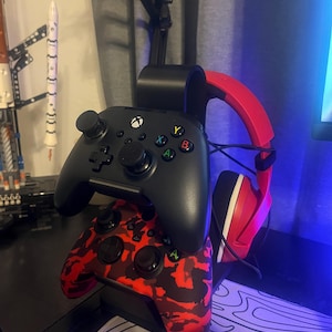 Op de afbeelding: Twee Xbox-controllers, één zwart en één rood camouflage, gestapeld op een zwarte standaard. Een rood-zwarte headset hangt rechts. Een modelraket is zichtbaar op de achtergrond.