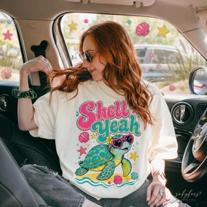 May include: Cream-colored t-shirt with the text "Shell Yeah" in pink and teal. The shirt features a cartoon turtle wearing heart-shaped sunglasses and a flower, surrounded by seashells and starfish.