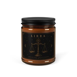 Peut inclure: Un pot de bougie en verre marron avec un couvercle noir. L'étiquette porte le mot "LIBRA" au-dessus d'une illustration d'une balance dorée, avec le texte "SEEK BALANCE, CHOOSE HARMONY" en dessous. La bougie est un article de décoration intérieure.