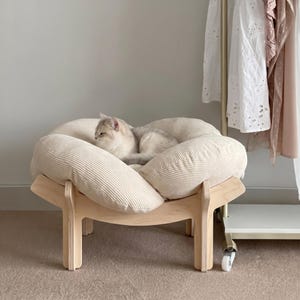Peut inclure: Un lit pour chat beige avec un chat clair à l'intérieur. Le lit a une texture semblable à du velours côtelé et est soutenu par un cadre en bois clair. Le lit est près d'un portant avec des vêtements blancs et roses.