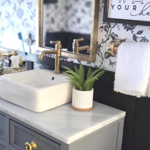 May include: Miniature bathroom scene with a white sink, gold faucet, and a small potted plant. The sink sits atop a gray cabinet with gold hardware. A mirror and towel rack complete the scene, with floral wallpaper in the background.