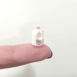 May include: Miniature white milk jug with a pink cap and label. The small plastic container has a handle and is designed to look like a real milk carton. The jug is resting on a finger.