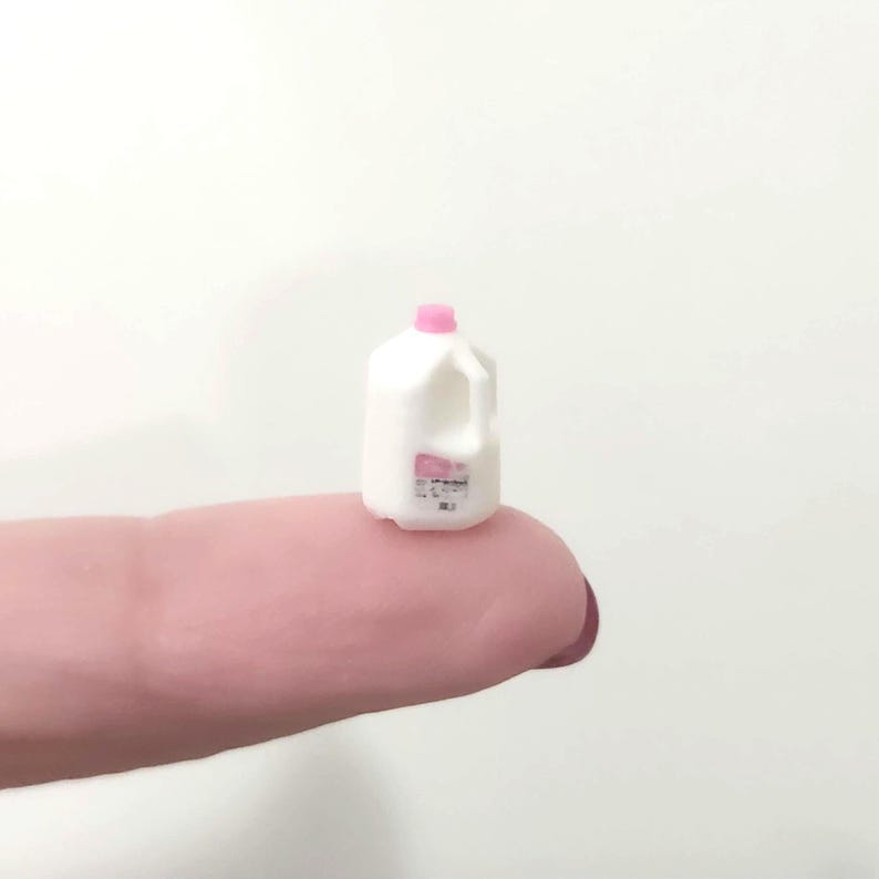 May include: Miniature white milk jug with a pink cap and label. The small plastic container has a handle and is designed to look like a real milk carton. The jug is resting on a finger.