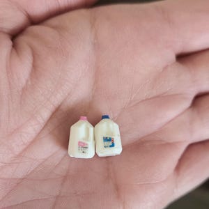 May include: Two miniature white plastic milk jugs, one with a pink cap and the other with a blue cap. Each jug has a label with text and a handle. The jugs are small enough to fit in the palm of a hand.