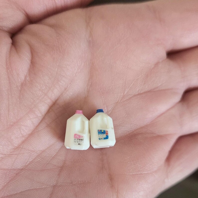 May include: Two miniature white plastic milk jugs, one with a pink cap and the other with a blue cap. Each jug has a label with text and a handle. The jugs are small enough to fit in the palm of a hand.
