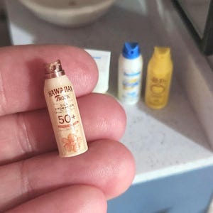 May include: Miniature sunscreen bottles, including a beige "Hawaiian Tropic" bottle with a floral design and "50+" text. Two other small bottles with blue and yellow caps are also visible. The items are small and appear to be dollhouse accessories.
