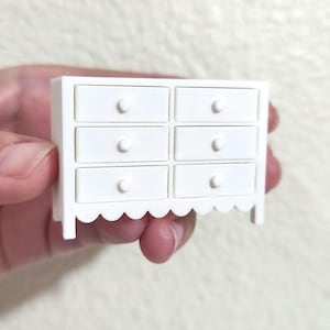 May include: A miniature white dresser with six drawers and a scalloped base. Each drawer has a small, rounded knob. The small furniture piece is held in a hand, suggesting its small size, and is likely for a dollhouse or similar project.